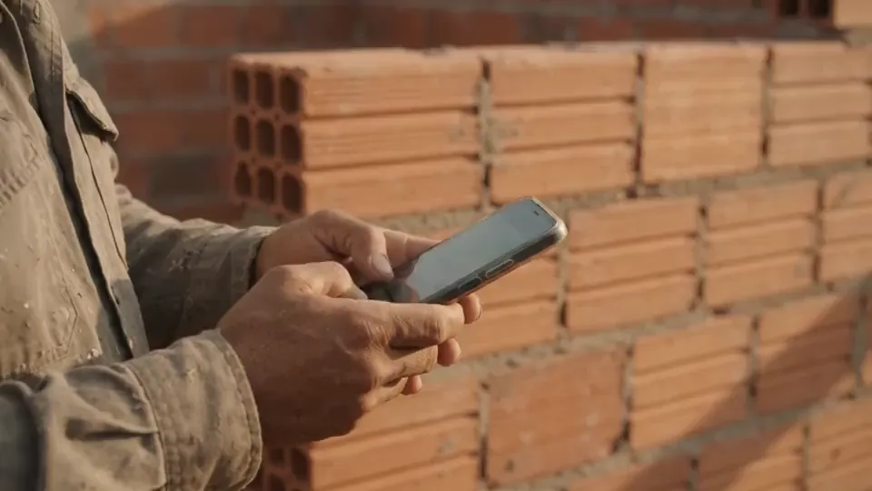 Mestre de obras pedindo orçamento pelo celular na frente da parede de tijolos